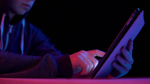 Un giovanotto col cappuccio con un tablet. L'hacker ha hackerato il tablet. Luce blu e rossa cade su un uomo su uno sfondo nero . — Foto Stock