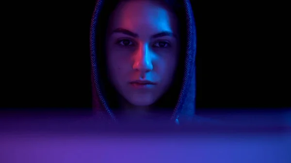 Young woman in a hood with a laptop. Hacker makes a hack through a laptop. Blue and red light falls on a woman on a black background. The camera goes down. — Stockfoto