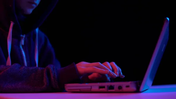 Young woman in a hood with a laptop. Hacker makes a hack through a laptop. Blue and red light falls on a woman on a black background. — Φωτογραφία Αρχείου