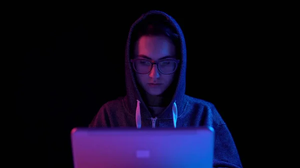 Jeune femme dans un capot avec un ordinateur portable. Un pirate pirate un ordinateur portable. La lumière bleue et rouge tombe sur une femme sur un fond noir. La caméra zoome sur la femme . — Photo