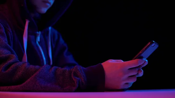 Giovane donna in un cappuccio con uno smartphone. L'hacker ha hackerato il telefono. Luce blu e rossa cade su una donna su uno sfondo nero . — Foto Stock