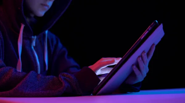 Giovane donna in cappuccio con un tablet. L'hacker ha hackerato il tablet. Luce blu e rossa cade su una donna su uno sfondo nero . — Foto Stock
