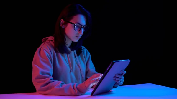 Uma jovem com um tablet. Uma mulher está a usar um tablet. Luz azul e vermelha cai sobre uma mulher em um fundo preto . — Fotografia de Stock