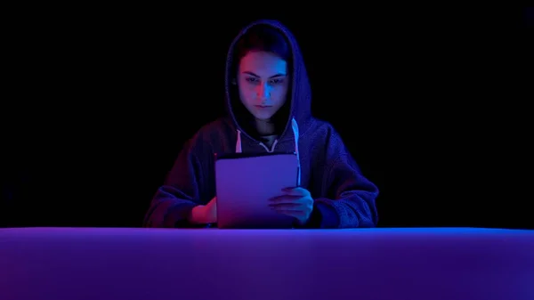 Young woman in a hood with a tablet. Hacker makes a hack through the tablet. Blue and red light falls on a woman on a black background. — Stock Fotó