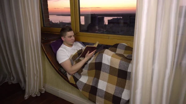 En ung man läser en bok. En man ligger på en fönsterbräda vid fönstret med en bok i händerna. Från kvällen. — Stockfoto