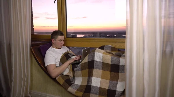 一个年轻人正在笔记本电脑上打字。一个人躺在窗台上，手里拿着笔记本电脑。晚上在外面 — 图库照片