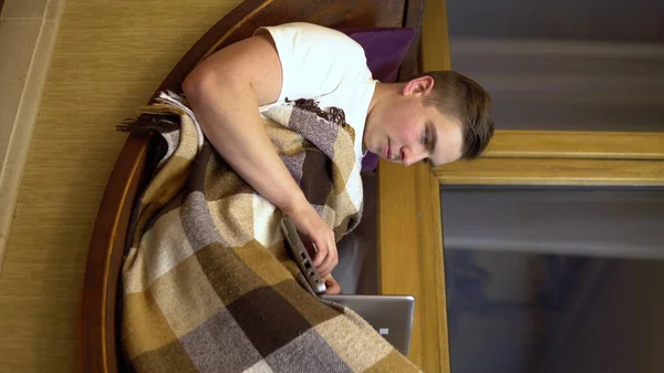 Um jovem está digitando em um laptop. Um homem deita-se numa soleira da janela com um portátil nas mãos. Fora da noite. Foto vertical . — Fotografia de Stock