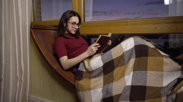 Uma jovem está lendo um livro. Uma menina se deita em uma soleira da janela junto à janela com um livro nas mãos. Fora da noite — Fotografia de Stock