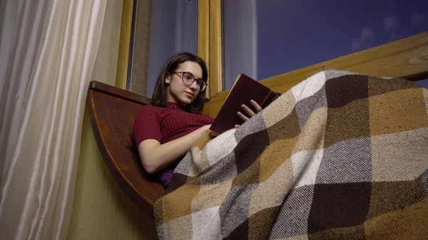 Uma jovem está lendo um livro. Uma menina se deita em uma soleira da janela junto à janela com um livro nas mãos. Fora da noite — Fotografia de Stock