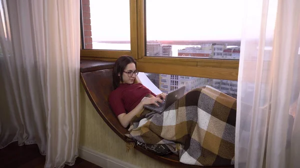Uma jovem está digitando em um laptop. Uma menina jaz em um peitoril da janela pela janela com um laptop em suas mãos. Do lado de fora da janela um dia brilhante . — Fotografia de Stock