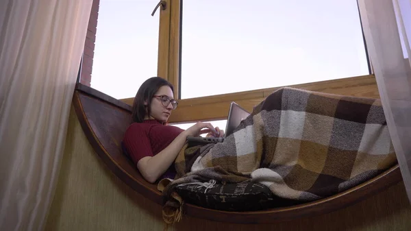 Une jeune femme tape sur un ordinateur portable. Une fille est allongée sur un rebord de fenêtre près de la fenêtre avec un ordinateur portable dans les mains. Devant la fenêtre un jour lumineux . — Photo