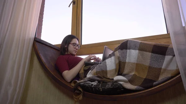 Uma jovem está digitando em um laptop. Uma menina jaz em um peitoril da janela pela janela com um laptop em suas mãos. Do lado de fora da janela um dia brilhante . — Fotografia de Stock