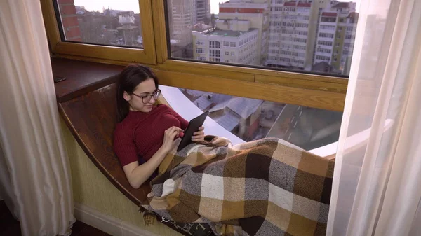 Uma jovem está a enviar mensagens no tablet. Uma menina está deitada em uma soleira da janela junto à janela com um tablet em suas mãos. Do lado de fora da janela um dia brilhante . — Fotografia de Stock