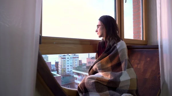 Una joven está sentada junto a la ventana. Una mujer se sienta en un alféizar de la ventana envuelta en una manta a cuadros y mira por la ventana. Fuera de la ventana un día brillante . — Foto de Stock