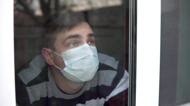 Ein trauriger junger Mann sitzt zu Hause in Quarantäne. Ein Mann schaut aus dem Fenster und legt seine Hand ans Glas. Blick hinter das Glas. — Stockvideo