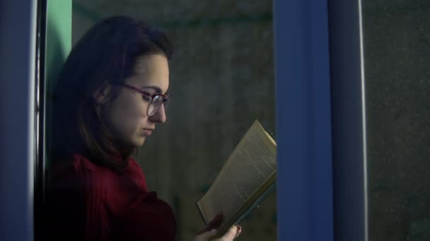 若い女性が本を手に家に座っている。少女は窓辺に座って本を読み、窓の外を見る。ガラスの後ろの景色. — ストック動画