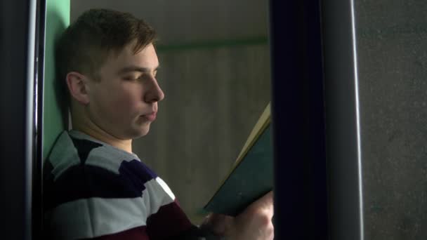 Mladý muž sedí doma s knihou v ruce. Muž sedí na parapetu, čte si knihu a pak se podívá z okna. Pohled za sklo. — Stock video