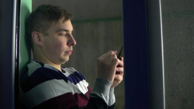 Genç bir adam elinde telefonuyla evde oturuyordu. Bir adam pencere eşiğinde oturur ve akıllı telefondan çalışır. Camın arkasına bak..