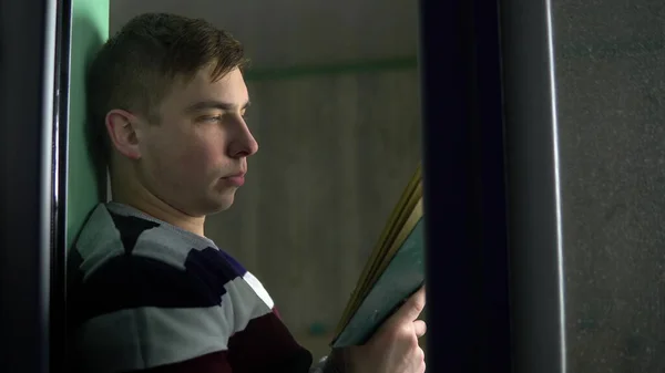 Un joven está sentado en casa con un libro en sus manos. Un hombre se sienta en un alféizar de la ventana y lee un libro y luego. Vista detrás del cristal . —  Fotos de Stock