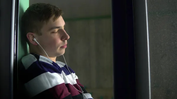 En ung man sitter hemma i karantän med hörlurar i öronen. En man sitter på ett fönsterbräde och lyssnar på musik. Utsikt bakom glaset. — Stockfoto