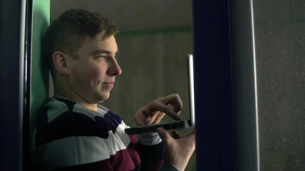 Un joven está sentado en casa con un portátil en sus manos. Un hombre se sienta en un alféizar de la ventana y trabaja a través de una computadora portátil. Vista detrás del cristal . —  Fotos de Stock