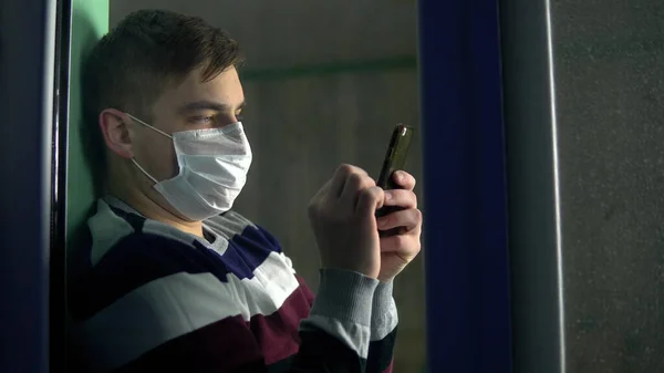 Ένας νεαρός άνδρας κάθεται στο σπίτι του σε καραντίνα με ένα τηλέφωνο στα χέρια του. Ένας άντρας κάθεται σε ένα περβάζι παραθύρου με μάσκα και δουλεύει μέσα από ένα smartphone. Προβολή πίσω από το γυαλί. — Φωτογραφία Αρχείου