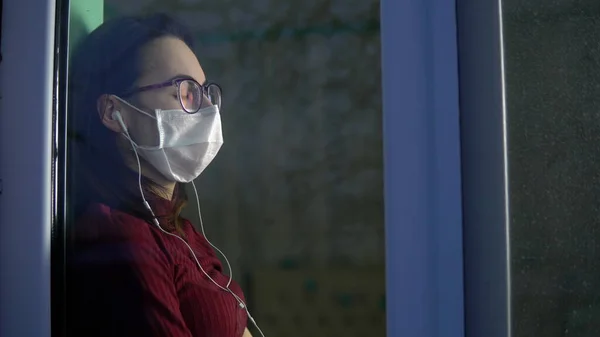 A young woman is sitting at home in quarantine with headphones in her ears. A girl sits on a window sill in a mask and listens to music. View behind the glass.