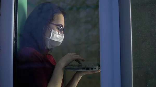 Una joven está sentada en su casa en cuarentena con un portátil en sus manos. Una chica se sienta en un alféizar de la ventana en una máscara y trabaja a través de un ordenador portátil. Vista detrás del cristal . — Foto de Stock