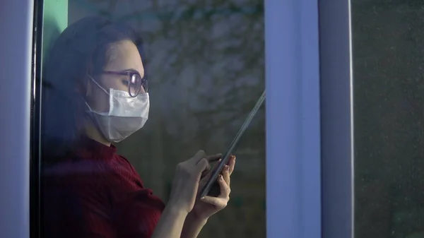 Una joven está sentada en su casa en cuarentena con una tableta en las manos. Una chica se sienta en un alféizar de la ventana en una máscara y funciona a través de un teléfono inteligente. Vista detrás del cristal . — Foto de Stock