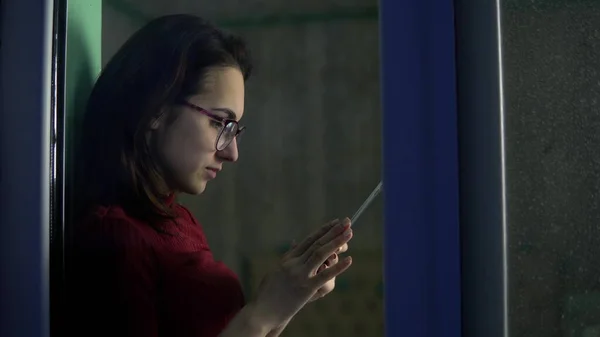 Egy fiatal nő ül otthon egy tablettával a kezében. Egy lány ül az ablakpárkányon, és okostelefonon keresztül dolgozik. Nézzen az üveg mögé.. — Stock Fotó