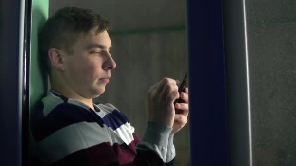 Un joven está sentado en casa con un teléfono en las manos. Un hombre se sienta en un alféizar de la ventana y trabaja a través de un teléfono inteligente y luego mira por la ventana. Vista detrás del cristal . — Vídeos de Stock