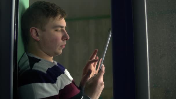 Młody mężczyzna siedzi w domu z tabliczką w rękach. Mężczyzna siedzi na parapecie, pracuje przez smartfona, a potem patrzy przez okno. Widok za szybą. — Wideo stockowe