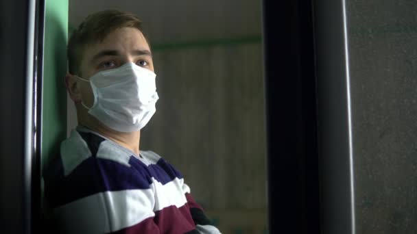 El joven triste está sentado en su casa en cuarentena. Un hombre se sienta en un alféizar de la ventana con una máscara y mira por la ventana. Vista detrás del cristal . — Vídeos de Stock