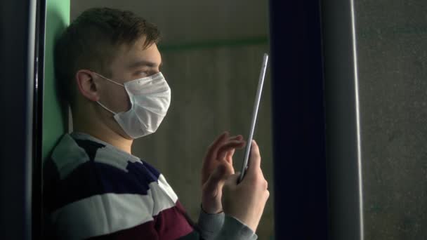 Un giovane uomo è seduto a casa in quarantena con una tavoletta in mano. Un uomo si siede su un davanzale con una maschera e lavora attraverso uno smartphone e poi guarda fuori dalla finestra. Vista dietro il vetro . — Video Stock