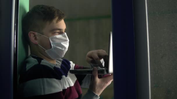 Ein junger Mann sitzt zu Hause in Quarantäne, einen Laptop in der Hand. Ein Mann sitzt maskiert auf einem Fensterbrett, arbeitet durch einen Laptop und blickt dann aus dem Fenster. Blick hinter das Glas. — Stockvideo