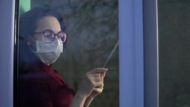 Genç bir kadın elinde tabletle karantinada oturuyor. Maskeli bir kız pencere pervazına oturur ve akıllı telefondan çalışır ve pencereden dışarı bakar. Camın arkasına bak..