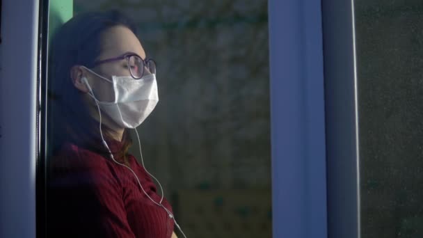 Una joven está sentada en su casa en cuarentena con auriculares en los oídos. Una chica se sienta en un alféizar de la ventana con una máscara y escucha música. Vista detrás del cristal . — Vídeos de Stock