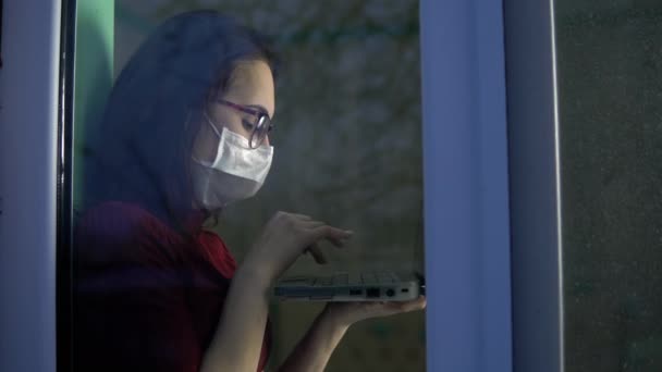 若い女性がノートパソコンを手に隔離されて家に座っている。少女は窓の上に座ってマスクをしてノートパソコンを通して作業し、窓の外を見る。ガラスの後ろの景色. — ストック動画