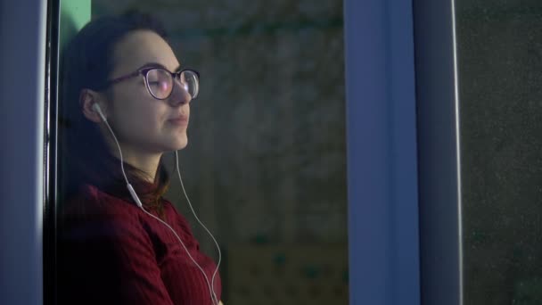 Una joven está sentada en su casa en cuarentena con auriculares en los oídos. Una chica se sienta en un alféizar de la ventana escucha música. Vista detrás del cristal . — Vídeos de Stock