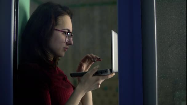 Una joven está sentada en casa con un portátil en sus manos. Una chica se sienta en un alféizar de la ventana y trabaja a través de una computadora portátil, y luego mira por la ventana. Vista detrás del cristal . — Vídeos de Stock