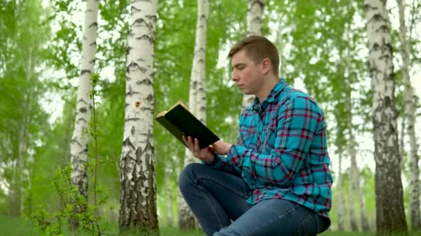 Egy fiatalember ül a természetben egy könyvvel a kezében. Egy férfi ül egy fatönkön a nyírfa erdőben, és könyvet olvas.. — Stock videók