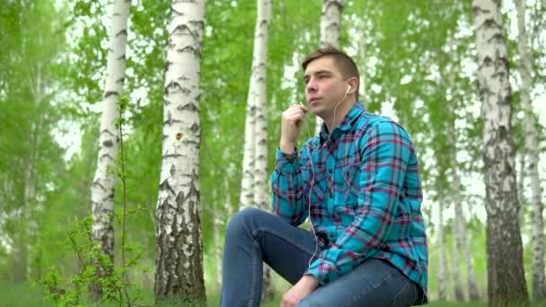 Ein junger Mann in der Natur telefoniert über Kopfhörer. Ein Mann sitzt mit Kopfhörern auf einem Baumstumpf in einem Birkenwald. — Stockvideo