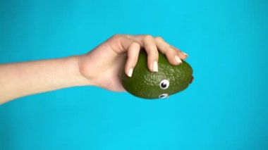 Bir kadının elinde gözleri olan bir avakado. Yaklaş. Avacado mavi bir arka planda etrafına bakıyor. Dikey video.