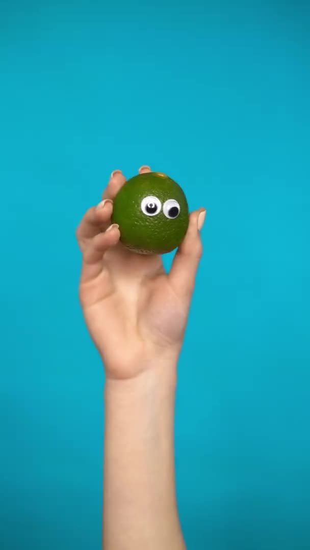 Lime with eyes in a woman hand close up. Lemon looks around on a blue background. Vertical video. — Stock Video