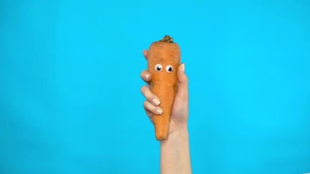 Carrot with eyes in a woman hand close-up. Carrot shakes and twists eyes on a blue background. Slow motion. — Stock Video