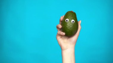 Bir kadının elinde gözleri olan bir avacado. Avacado çerçeveye atlıyor ve mavi arka planda etrafına bakıyor. Kadın eli yakın çekim.