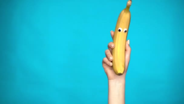 Μπανάνα με μάτια σε γυναικείο χέρι. Μπανάνα πηδά στο πλαίσιο και κοιτάζει γύρω σε ένα μπλε φόντο. Γυναικείο κοντινό πλάνο. — Αρχείο Βίντεο