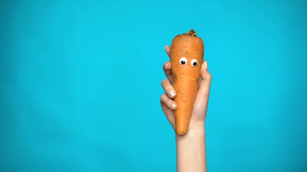 Carrot with eyes in a woman hand. Carrots jumps into the frame and looks around on a blue background. Woman hand close-up. — Stock Video