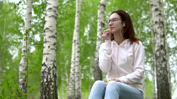 Una joven en la naturaleza está hablando por teléfono a través de auriculares. Una chica se sienta en un tocón en un bosque de abedules con auriculares . — Vídeos de Stock