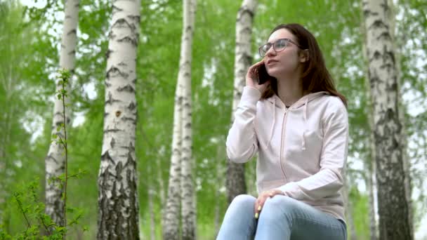 Jovem mulher na natureza com um telefone. Uma menina senta-se em um toco em uma floresta de bétula e fala ao telefone . — Vídeo de Stock